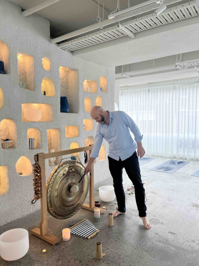 A man strikes a large gong in a modern, softly lit room with yoga mats and candles.