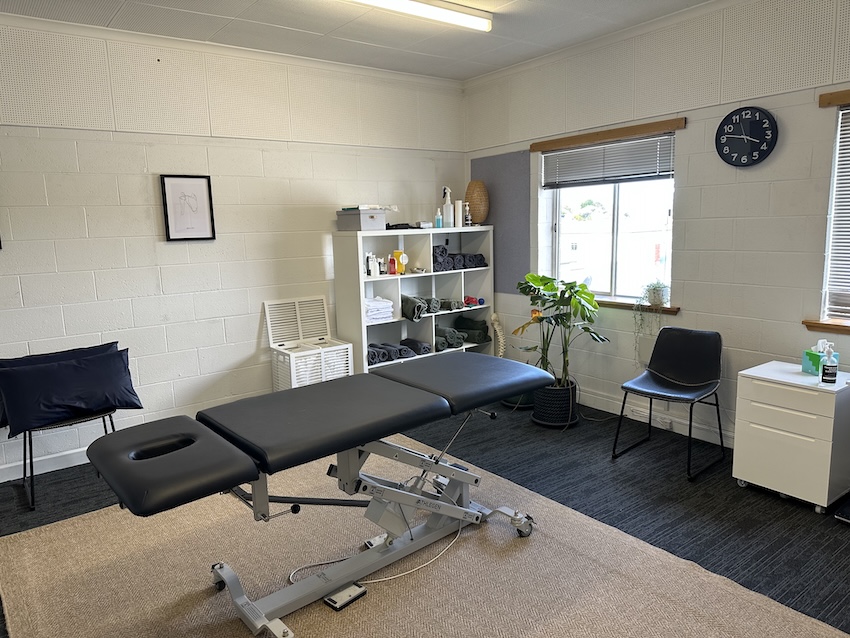 A treatment room in Acupuncture Hobart with an exam table, chairs, shelves, and a wall clock.