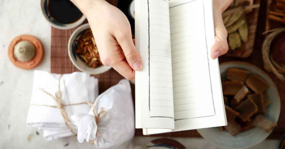 Hands holding an open notebook above a table with packages, bowls, and Herb-Drug Interactions notes.